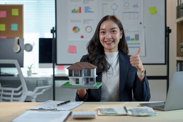 Asian saleswoman, real estate agent holding model house and writing lease contract, insurance, sales agreement, legal in collateral while sitting at desk in real estate project office.