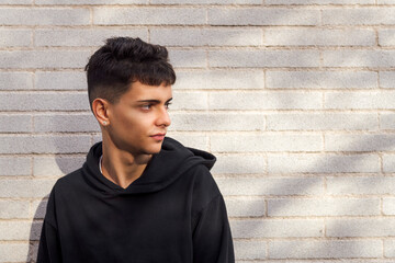 caucasian male teen wearing casual clothes standing in sunlight against white brick wall looking to the side, concept of youth and urban lifestyle, copy space for text