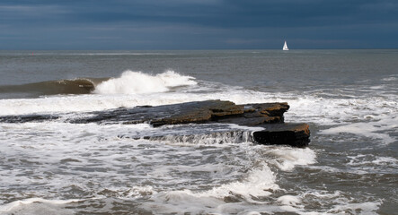 Waves breaking over coastal rocks with small sailing yacht on horizon