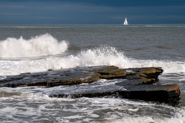 Waves breaking over coastal rocks with small sailing yacht on horizon