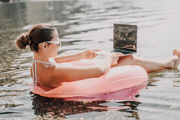 A woman is sitting in a pink inflatable raft on a lake, using a laptop. Concept of relaxation and...