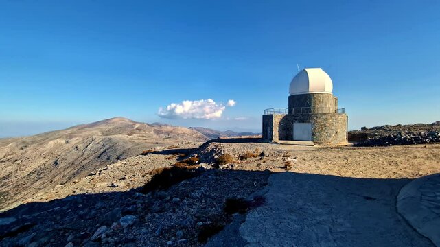 Scenic view of Skinakas Observatory perched on a rocky mountain, surrounded by rugged terrain and a bright blue sky in Crete, Greece