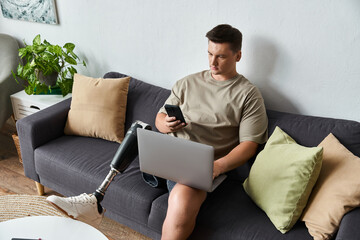 Handsome young man with a prosthetic leg sitting on a sofa, focused on his laptop and smartphone.