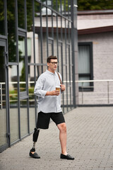 Handsome young man with prosthetic leg strolls outside, holding coffee and wearing casual attire.