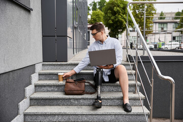 A handsome young man with a prosthetic leg is working on his laptop on outdoor steps.
