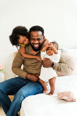 A joyful father sits on a bed, playfully holding his baby while his daughter hugs him, creating a...