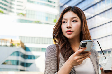 A young woman with long hair is holding a smartphone, looking thoughtfully ahead, with a drink in her other hand, set against a modern urban backdrop. Young businesswoman or entrepreneur concept.