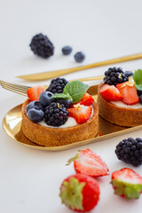 Tartlets with Fresh Blueberries, Strawberries, and Blackberries on a Gold Stand with Golden Cutlery on a White Background