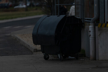 A black trash can is parked quietly on the side of the road, casting a shadow in the darkness of the night around it