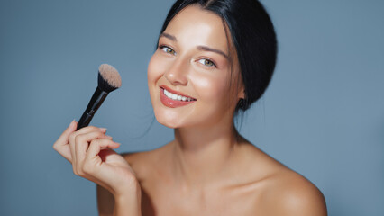 Portrait of a smiling woman holding a makeup brush, highlighting her radiant skin against a blue...