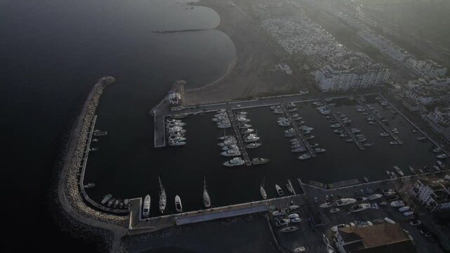 "Drone footage at sunset over La Duquesa Port in Sabinillas, M&aacute;laga. A bird's-eye view advances over the marina, showcasing docked boats, the harbor, and the serene coastal charm."