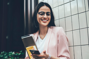 Half length portrait of cheerful influence blogger with modern cellphone device and textbook...