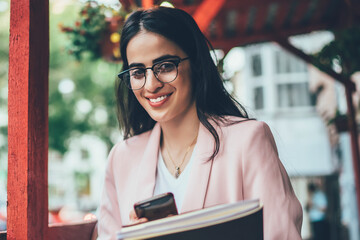 Half length portrait of cheerful influence blogger with modern cellphone device and textbook...