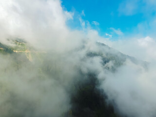 Winding mountain road disappears into clouds, with lush green forest below and blue sky peeking through, creating a mysterious and captivating scene