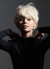 Elegant blonde woman with short hair, wearing a black top, poses with confidence against a dark background, exuding a sense of allure and sophistication.