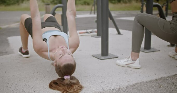 Two young women enjoy their workout in a park on a summer day. They perform various exercises like inverted rows, squats, and stretching.