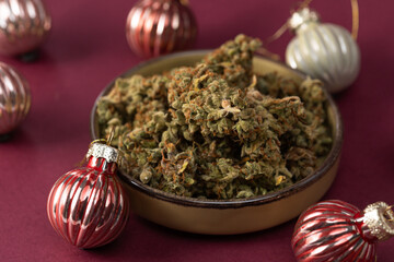 on a dark red background in a bowl some dry buds of medical marijuana, next to Christmas balls