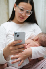 Beautiful young woman wearing white clothes is breastfeeding her baby at home, close-up back view. She is working online at the same time, remote work from home.