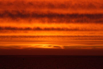 Dramatic sunset over a calm sea.  Fiery orange clouds streak the sky. Dark water reflects the...