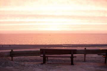 Tranquil beach scene at sunset.  Peaceful, muted colors of a pink-tinged sunrise over the water.  Wooden bench on the sand.