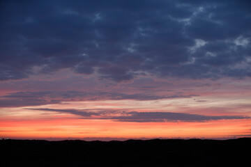 Fototapeta premium Sunset hues paint a dramatic sky above dark silhouettes of hills. Clouds transition from dark to vibrant orange-pink.