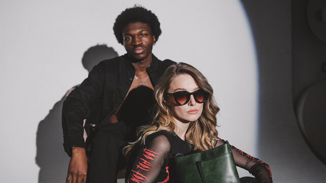 A diverse and fashionable couple poses against a white backdrop. The man wears a black open shirt, and the woman has sunglasses, a black mesh top with red designs, and holds a dark green handbag. 