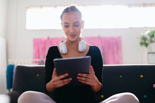 A young ballerina sits cross-legged on a gray couch, wearing white headphones and using a tablet, with a pink curtain and vibrant modern decor visible in the background.