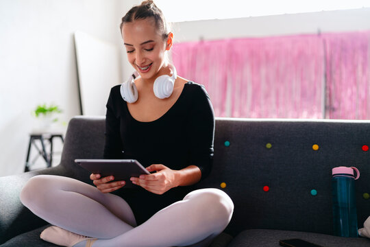 A young ballerina sits cross-legged on a gray couch, wearing white headphones and using a tablet, with a pink curtain and vibrant modern decor visible in the background.