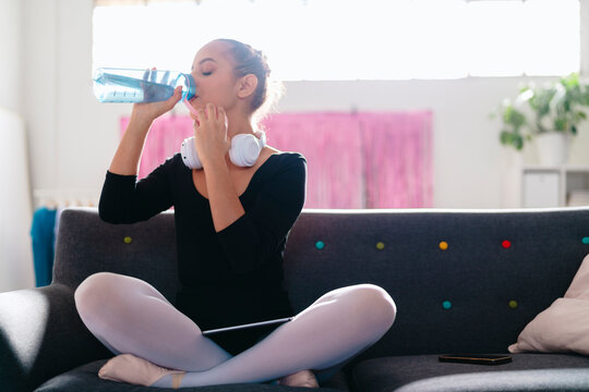 A young ballerina sits cross-legged on a gray couch, wearing headphones and drinking water from a blue bottle, with a bright modern room and pink curtain in the background.