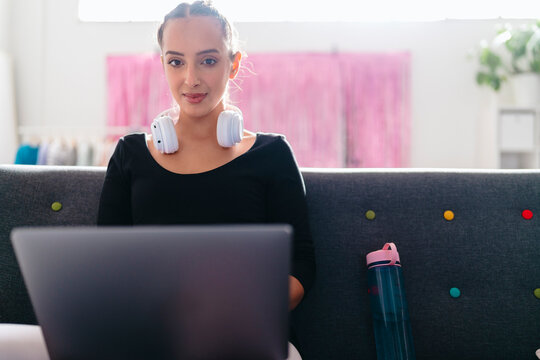 A young ballerina with headphones around her neck uses a laptop while sitting on a gray couch, with a bright studio environment featuring pink curtains and greenery in the background.