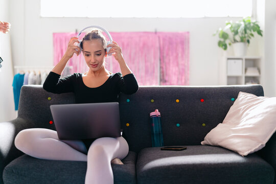 A young ballerina sits on a gray couch, wearing white headphones and interacting with a laptop, in a bright modern studio featuring pink curtains and vibrant decor.
