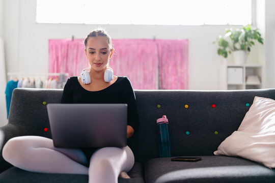 A young ballerina with headphones around her neck uses a laptop while sitting on a gray couch, with a bright studio environment featuring pink curtains and greenery in the background.