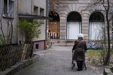 An elderly woman wearing a brown quilted coat and white knitted hat uses a walker while strolling down a narrow path in an urban residential area