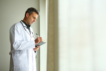 A male doctor standing by the window of his private office reviewing reports