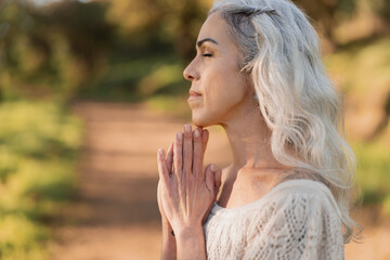 A mature woman with gray hair meditates peacefully outdoors in a natural setting during sunset. The calm and tranquil scene highlights connection with nature and personal mindfulness.