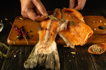 Hands skillfully place fresh herbs on a fillet of salmon lying on a wooden cutting board. Red chilies and spices surround the fish, enhancing its vibrant colors and flavors