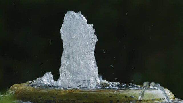 Flow of clean water is splashing out upwards from a small artificial water fountain with blurry natural background.
