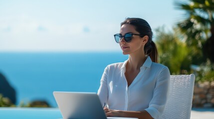 A focused woman works on her laptop by the pool, enjoying a beautiful view of the ocean on a sunny day.