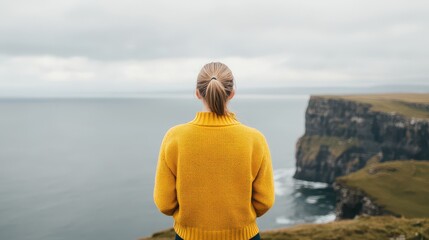 Fototapeta premium A person in a yellow sweater stands by the cliffs, gazing at the tranquil ocean, surrounded by dramatic coastal scenery.