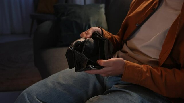 Young man spending evening at home sitting on sofa in living room, opens wallet and looks inside for cash, shaking wallet no money, side shot.