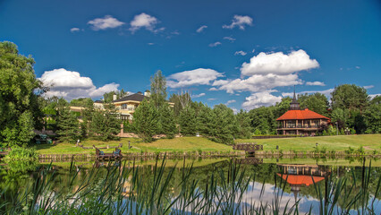 Fototapeta premium View of the pond, blooming meadow, forest and cottages in village at summer timelapse hyperlapse