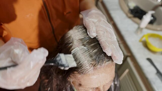 Hairdresser applying dye to a senior woman's gray hair, using a brush and wearing gloves for a professional touch