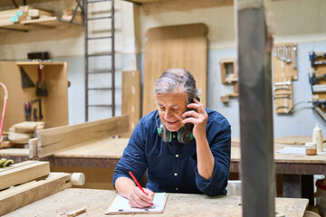 Mature craftsman in a lumberyard taking notes on a phone call