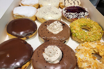 A box of assorted gourmet donuts featuring a variety of toppings and fillings, including chocolate glaze, powdered sugar, vanilla frosting, crushed nuts, and whipped cream.