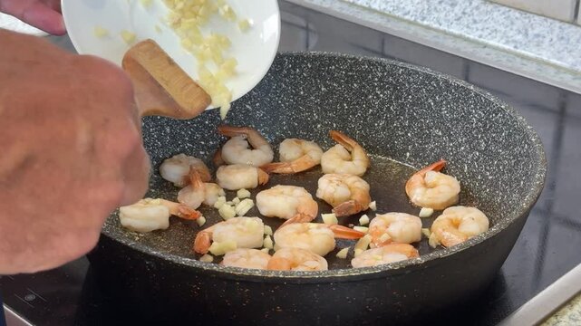 A person adds minced garlic to sizzling shrimp in a skillet while preparing a delicious meal in the kitchen.