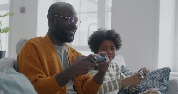 African American man playing video game with cute little son having fun with child in apartment. Family lifestyle and gaming concept.