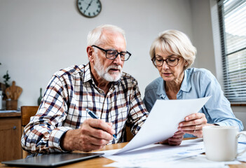 Elderly couple reviewing financial documents together at home while discussing plans. Laptop and coffee signify a modern, comfortable, and collaborative environment