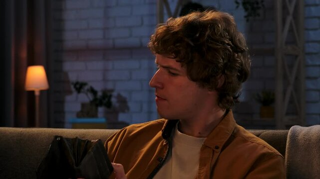 Young man spending evening at home sitting on sofa in living room, holding wallet looking for money inside, worried expression, shaking empty wallet, close up.