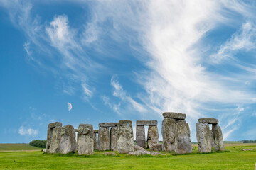 Moon in blue sky with feather clouds above Stonehenge prehistoric structure on Salisbury Plain, England consisting of outer ring of sarsen standing stones and inside ring of smaller bluestones