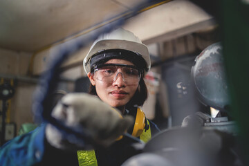 A woman wearing a hard hat and safety glasses is looking at a control panel. She is wearing a yellow vest and is wearing a pair of gloves